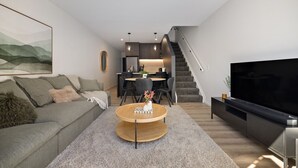 A modern living room with a sofa, coffee table, TV, and a staircase leading to a kitchen area. - Prime Location with mountain views (Queenstown)