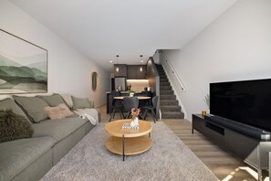A modern living room with a sofa, coffee table, TV, and a staircase leading to a kitchen area. - Prime Location with mountain views (Queenstown)