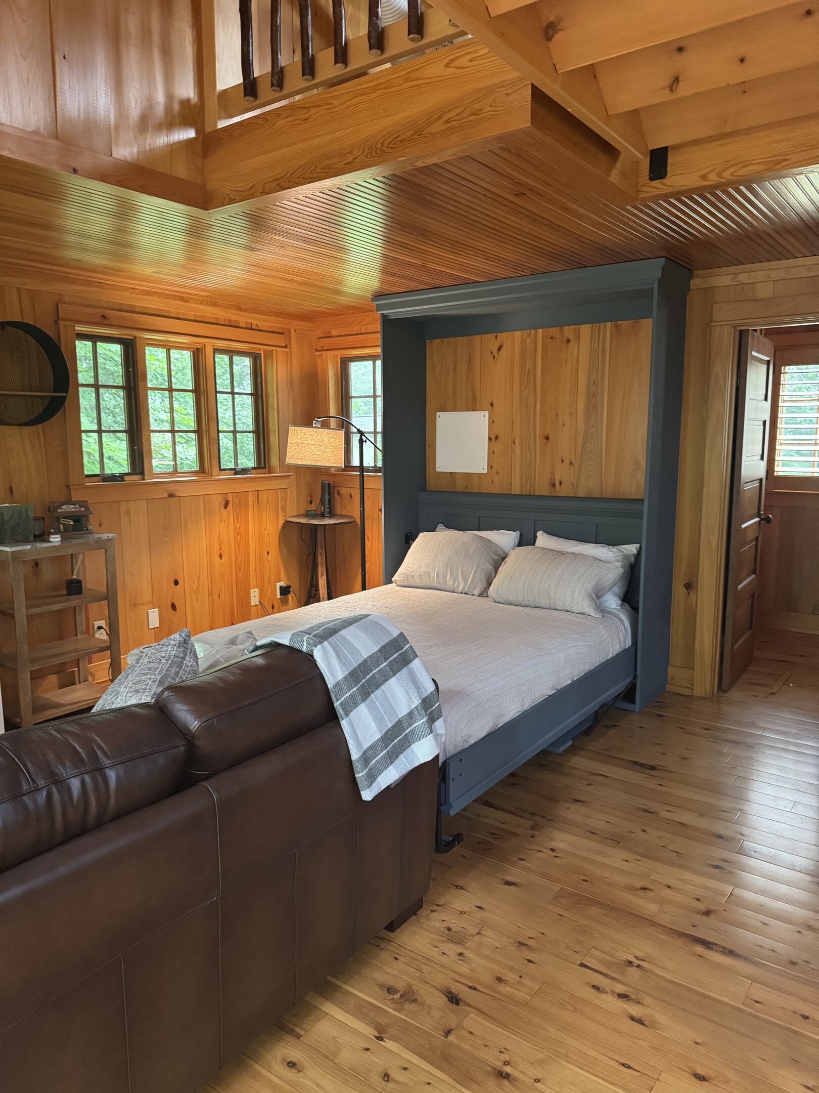 A wooden cabin interior with a bed, a brown leather couch, a lamp, and a window with a view of trees.