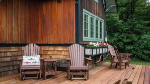 A wooden deck with Adirondack chairs and a table, a wooden building with green windows, and a flower bed with red and white flowers. - Magical Jordan River Oasis | Private Cabin & Deck (East Jordan)