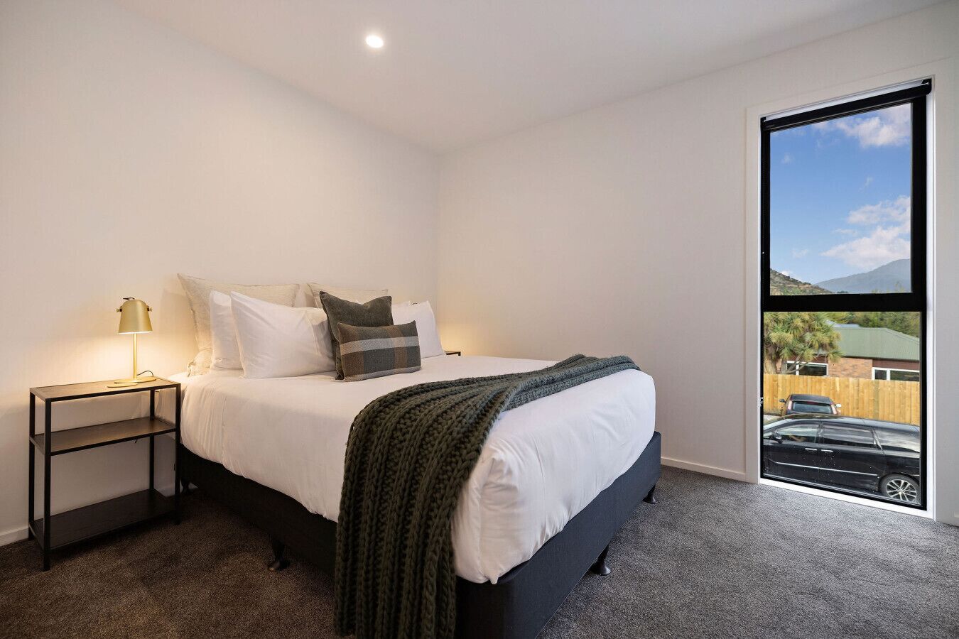 A bedroom with a bed, a nightstand, a lamp, and a view of a car and mountains through a large window.