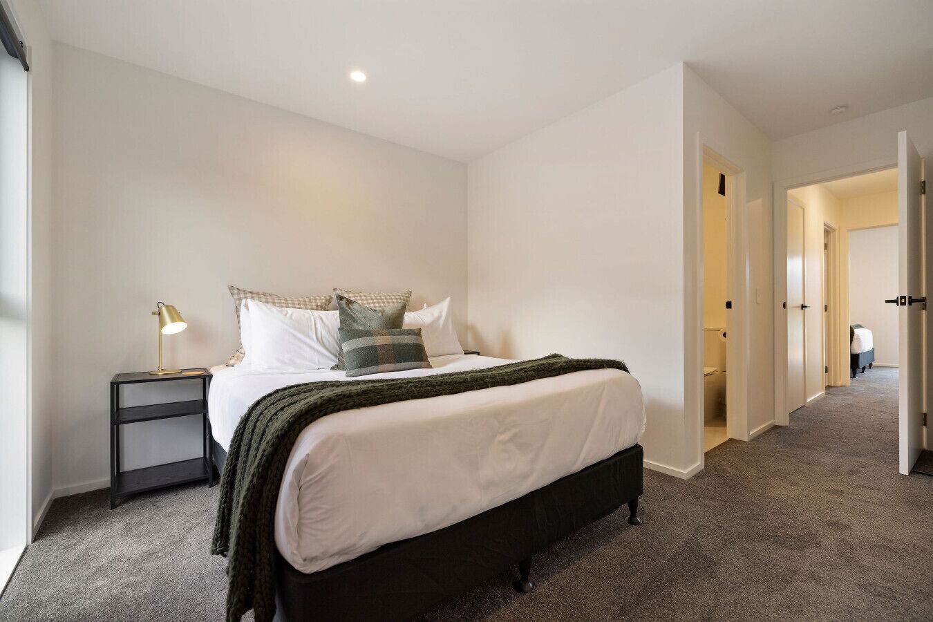 A bedroom with a bed, bedside tables, and a lamp. There is a hallway leading to other rooms.