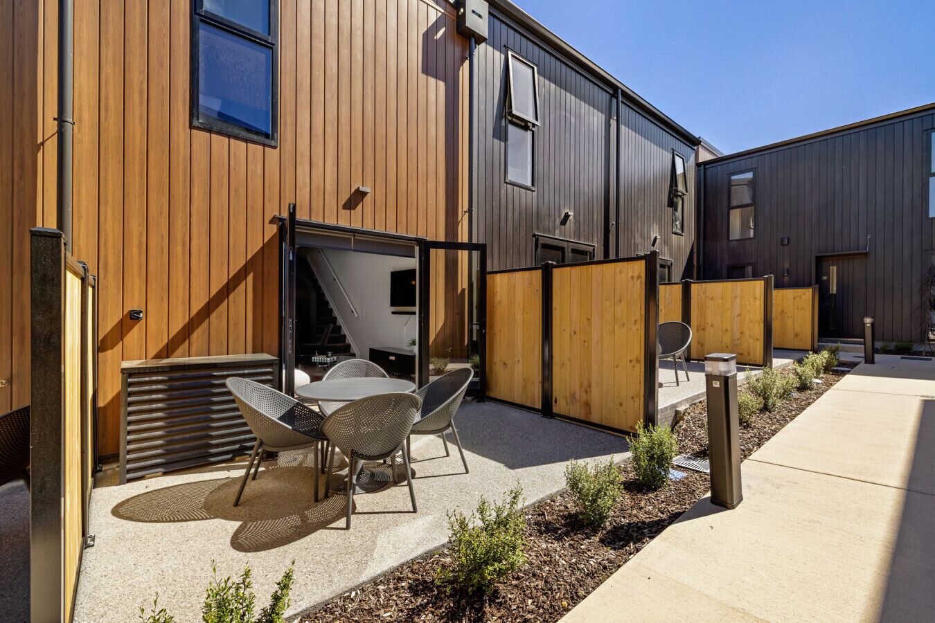 A modern building with a wooden exterior, a covered outdoor seating area with chairs and a table, and a clear sky.