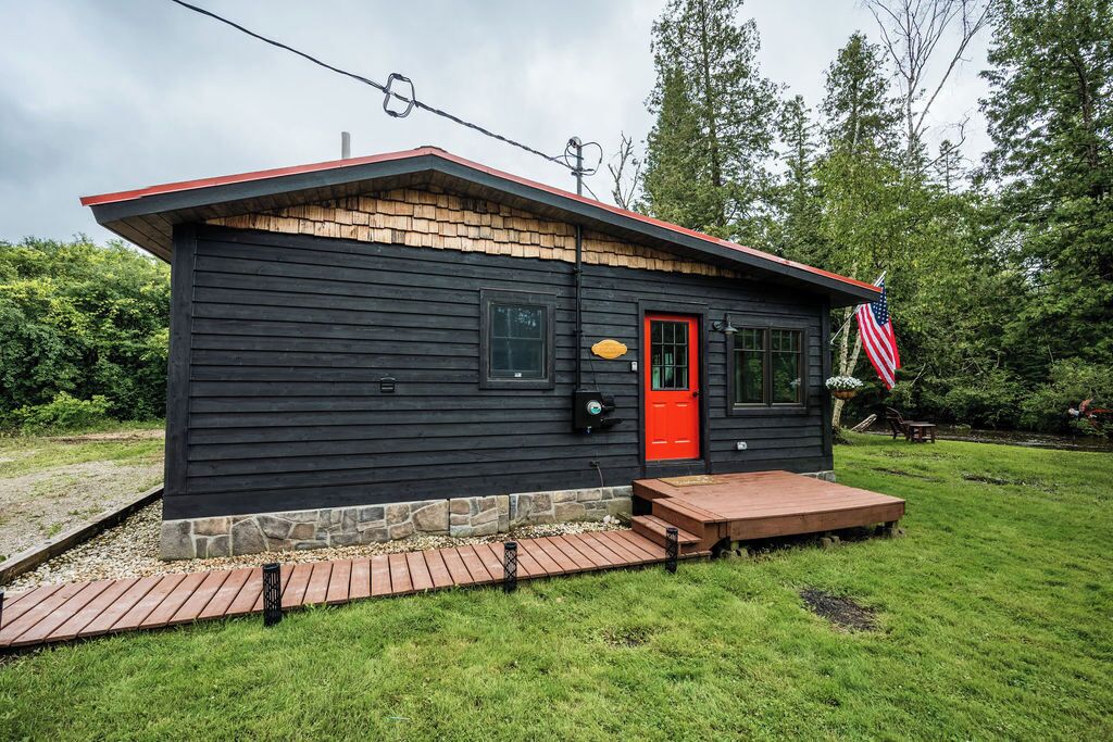 Una cabaña pequeña con puerta roja, terraza de madera y una bandera estadounidense.