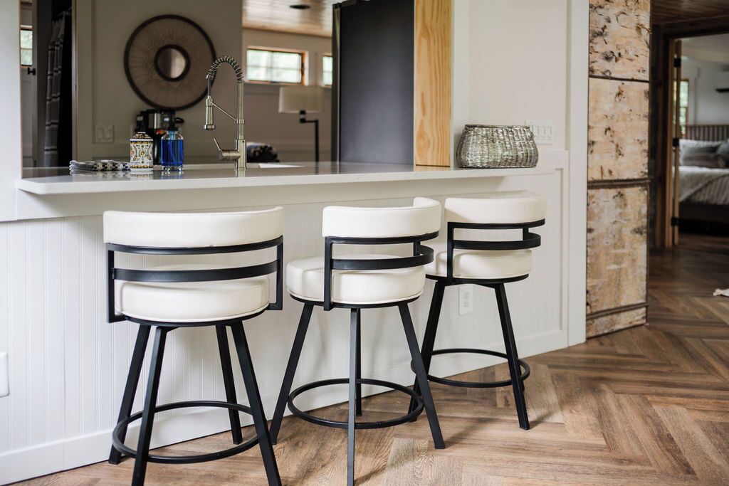 Une cuisine avec un plan de travail blanc, trois tabourets de bar et un plancher en bois.