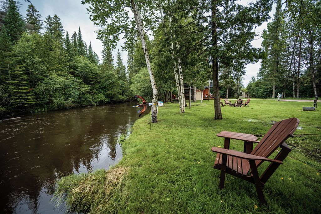 A riverside setting with a wooden chair and a small table, surrounded by trees and a grassy area.