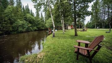 A riverside setting with a wooden chair and a small table, surrounded by trees and a grassy area.