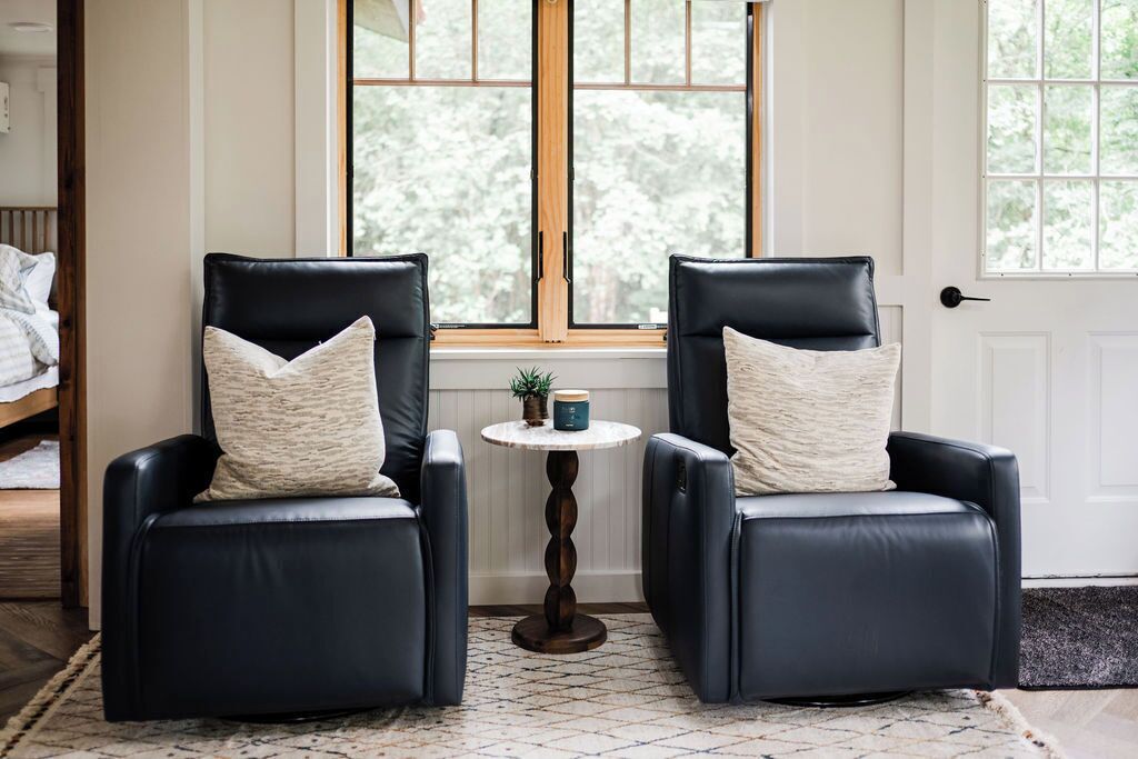 Two black leather chairs with matching cushions, a small round table with a plant and a mug, and a window with a view of trees.