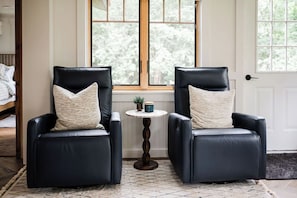Two black leather chairs with matching cushions, a small round table with a plant and a mug, and a window with a view of trees.