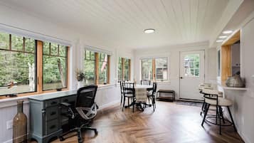 Una habitación con piso de madera, un escritorio con una silla, una mesa de comedor con sillas y ventanas amplias con vista a árboles.