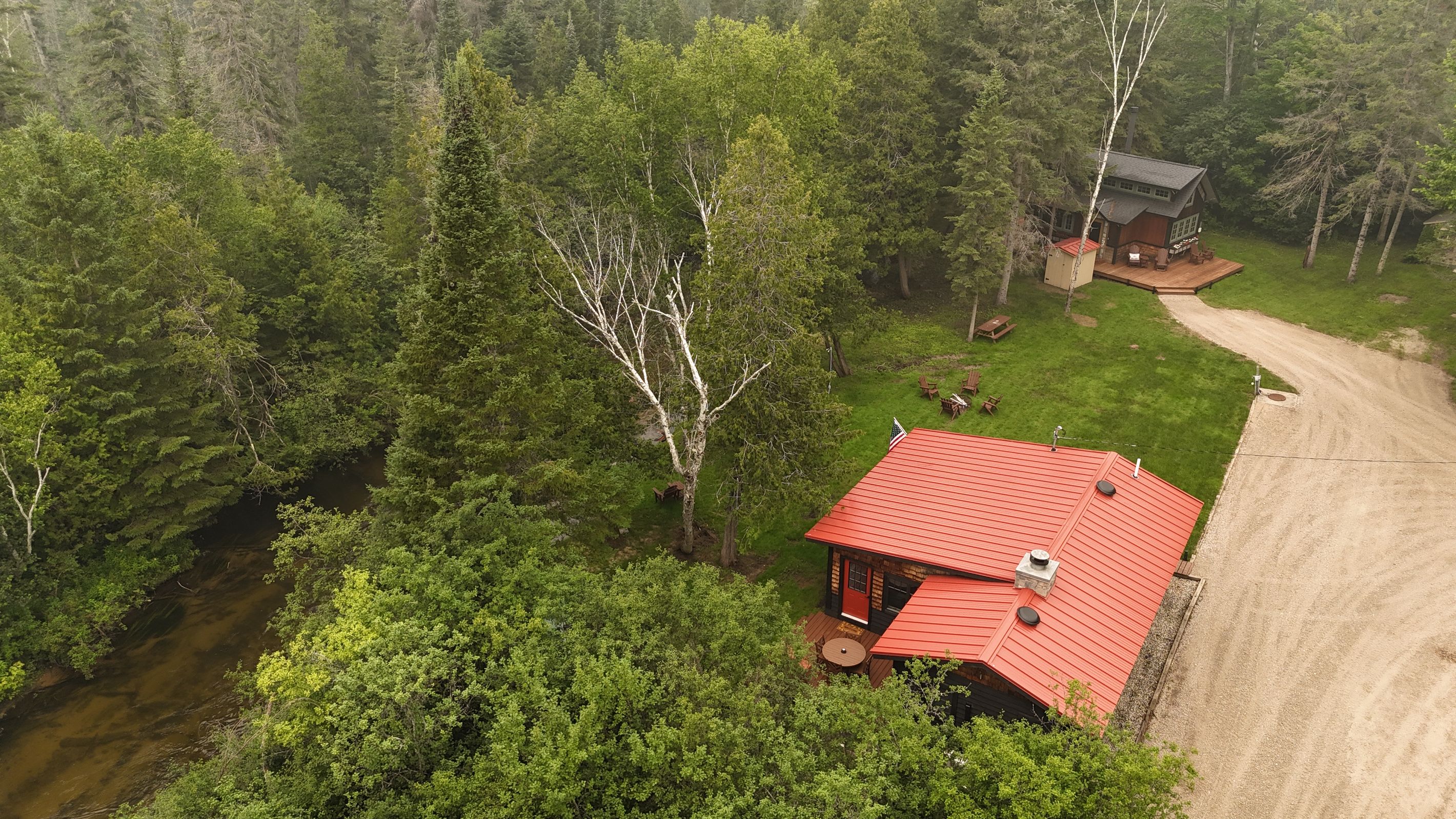 A cabin with a red roof surrounded by dense forest, a dirt path leading to it, and a small deck with outdoor seating.