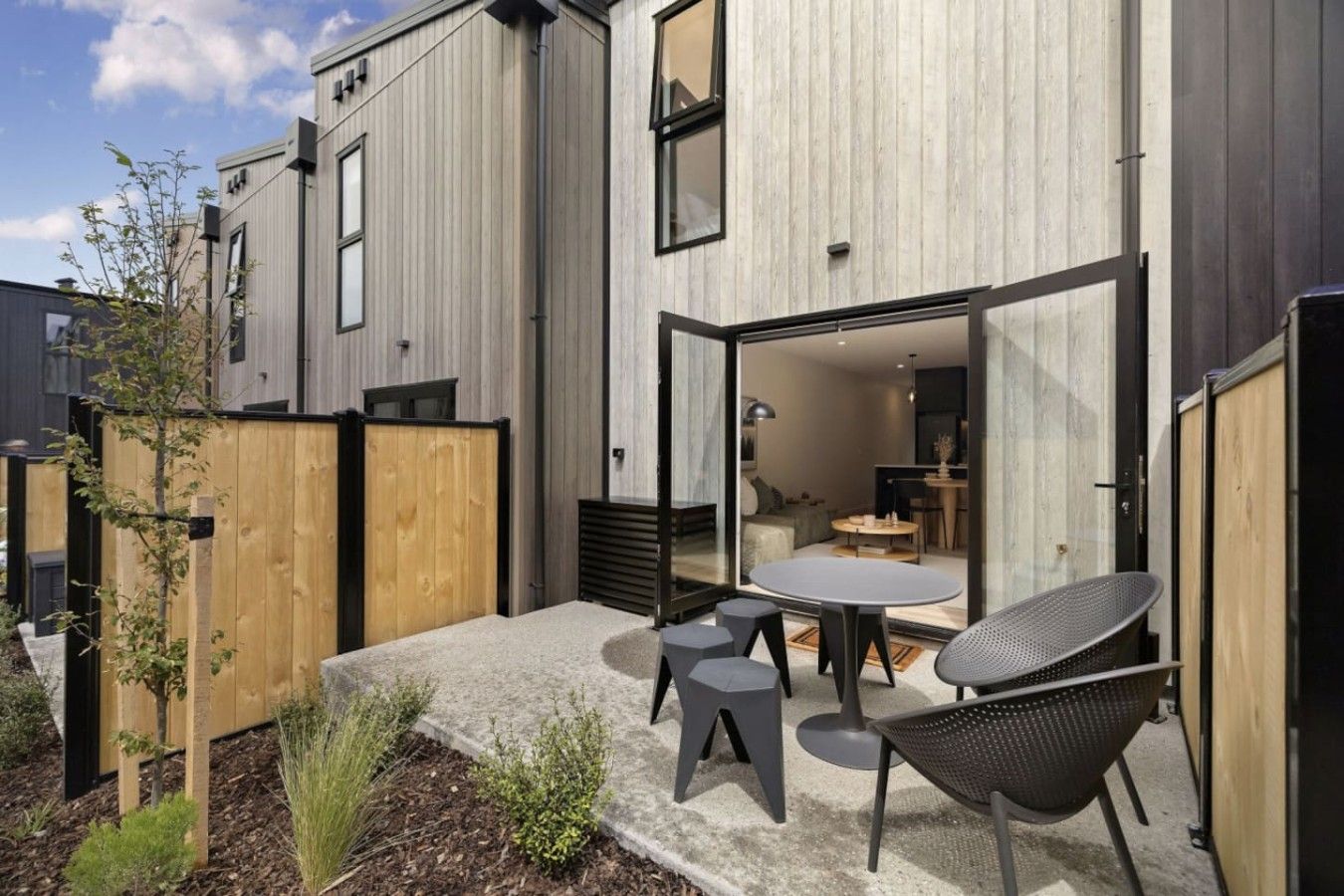 A modern building with a patio area featuring a table and chairs, a wooden fence, and a small garden.