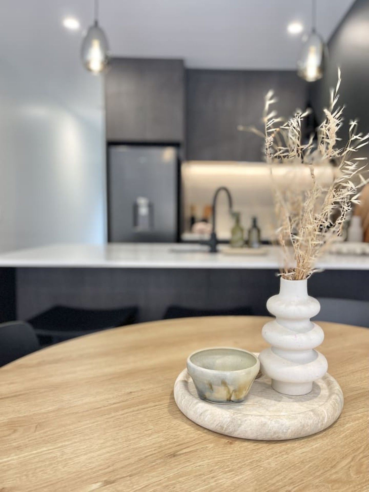 Cozinha moderna com ilha central, luminárias pendentes e um vaso decorativo com plantas secas.