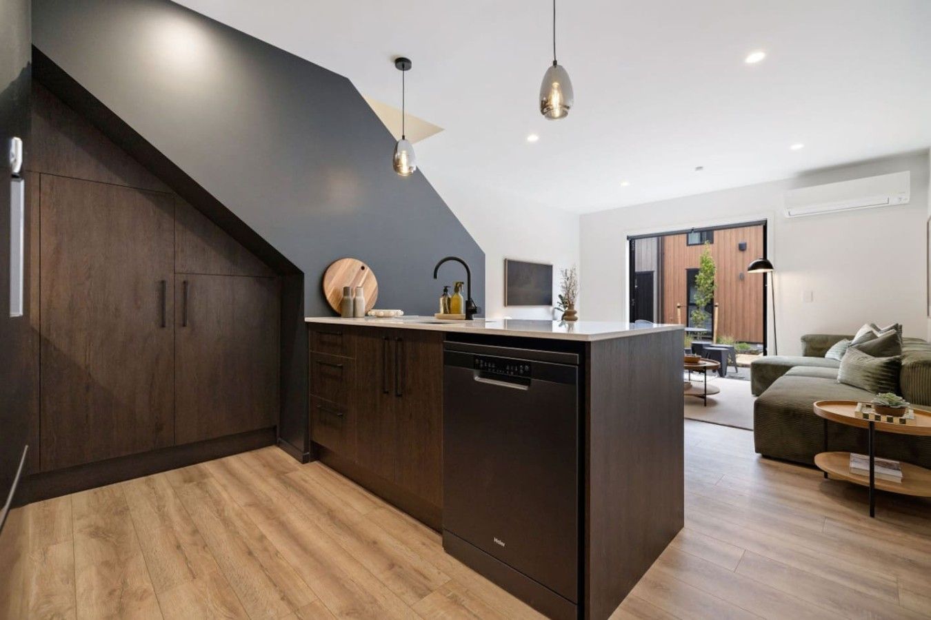A modern kitchen with dark cabinetry, a central island, and a dishwasher. There is a dining area with a sofa and a table.