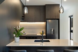 A modern kitchen with dark wood cabinets, a stainless steel refrigerator, pendant lighting, and a central island with a sink and bar stools. - Prime Location with mountain views (Queenstown)