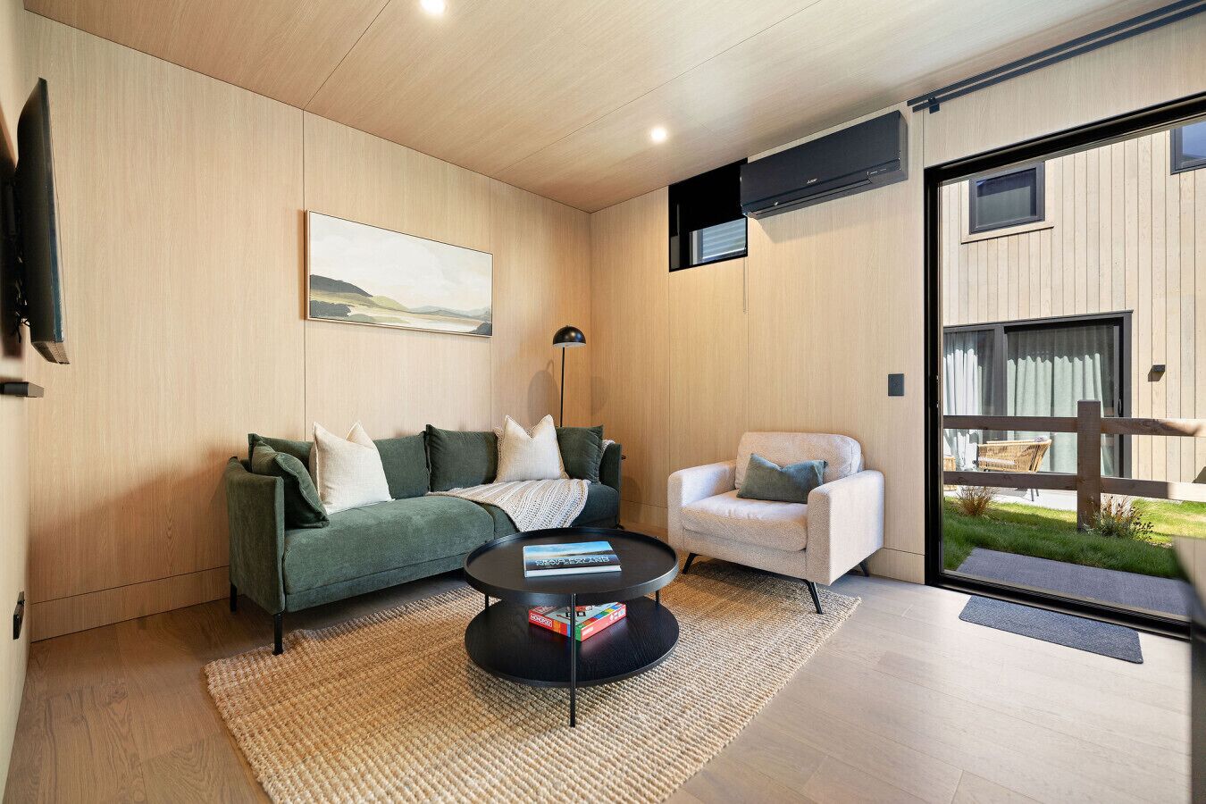 Een moderne woonkamer met een groene bank, een witte fauteuil, een salontafel en een groot raam met uitzicht op de omgeving.