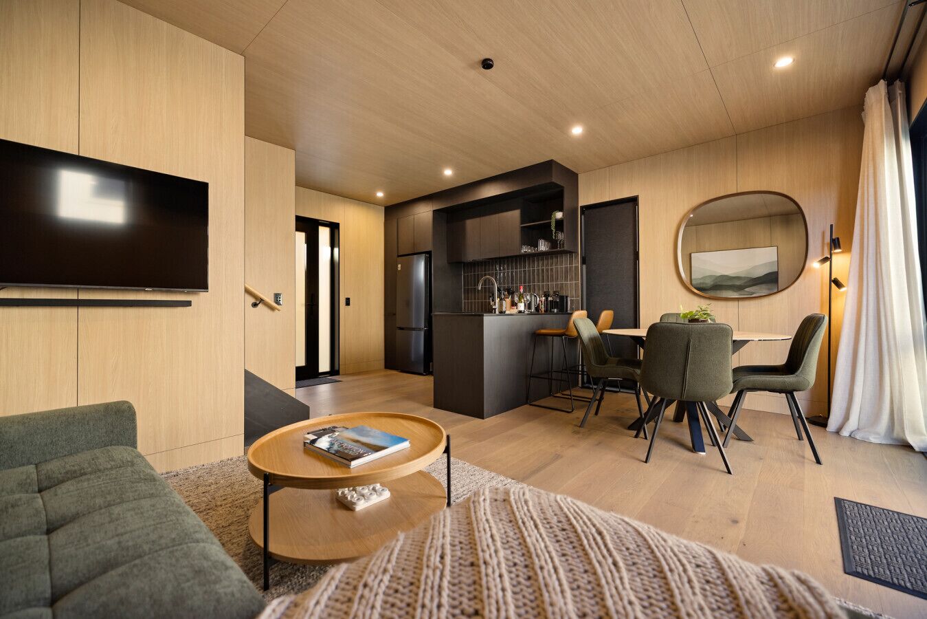 A modern living room with a flat-screen TV, a sofa, a coffee table, and a dining area with chairs and a small kitchen.