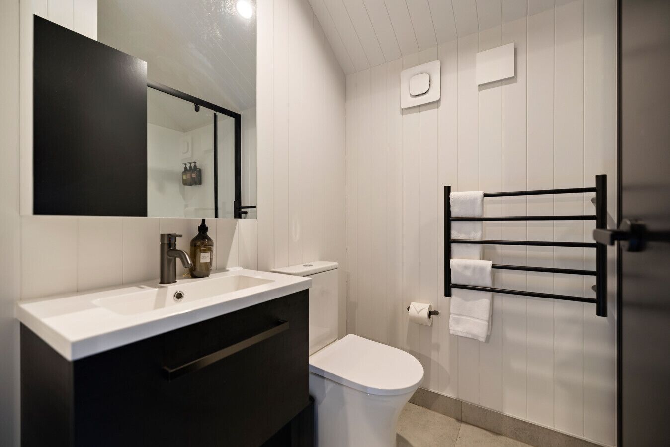 A modern bathroom with a white sink, dark vanity, a toilet, a towel rack, and a mirror.