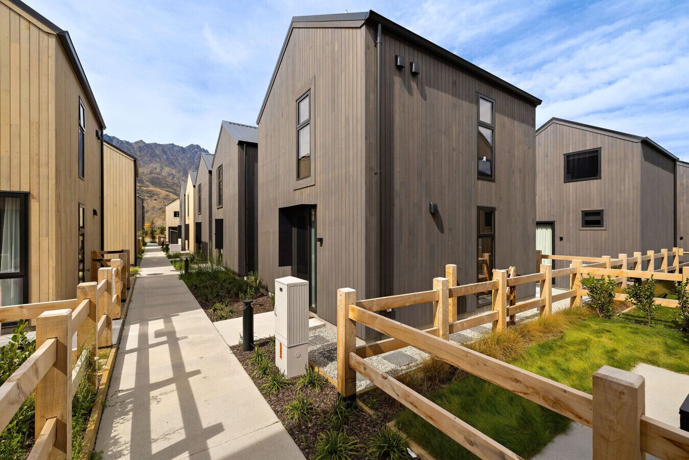 A row of modern buildings with a wooden fence in front, a paved walkway, and a mountainous backdrop.