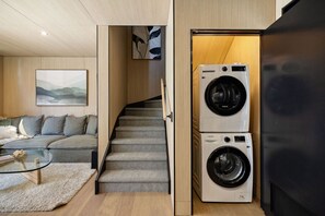 Ein kompakter Wohnbereich mit Sofa, Glastisch, Treppe und übereinander gestapelten Waschmaschinen.