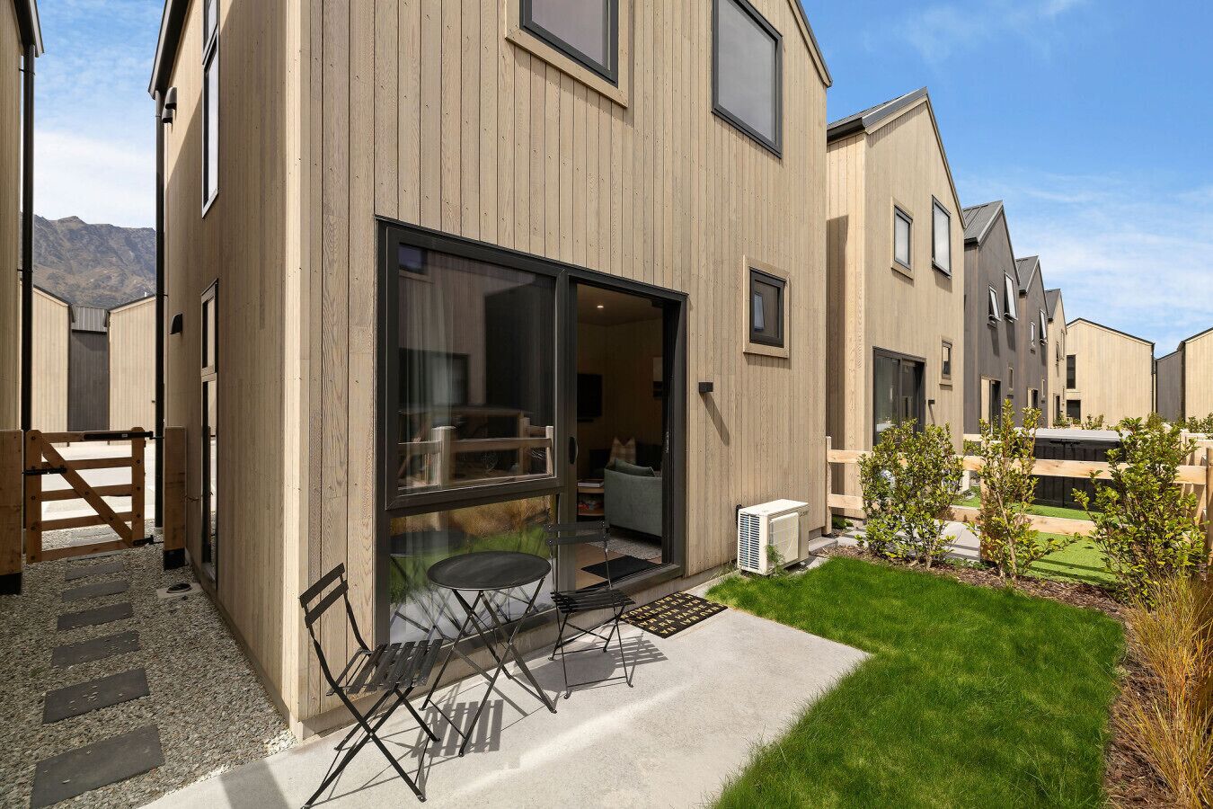 A modern building with a sliding glass door entrance, a small patio with a table and chairs, and a well-maintained lawn.