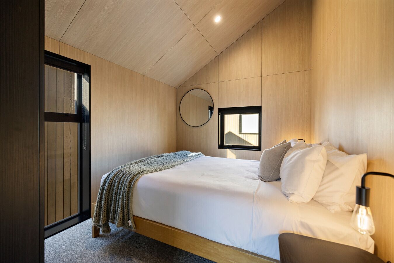 A bedroom with a bed, a bedside lamp, a window, and a mirror on the wall.