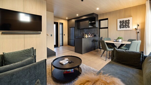A modern living room with a sofa, a coffee table, and a TV. There is a kitchen area with a refrigerator and a dining table with chairs. - Luxury Queenstown 2Bed Villa (Queenstown)