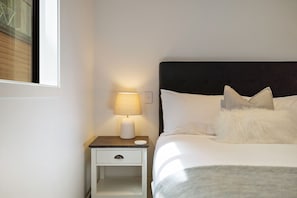 Ein ordentlich bezogenes Bett mit weißer Bettwäsche und zwei Kissen, eine Nachttischlampe auf einem kleinen weißen Nachttisch und ein Fenster mit teilweise geöffneten Jalousien.