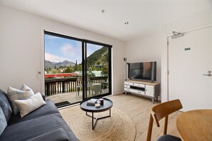 Ein modernes Wohnzimmer mit einer großen Schiebeglastür, die auf einen Balkon führt, einem Flachbildfernseher, einem Sofa, einem Couchtisch und einem Holzsessel.
