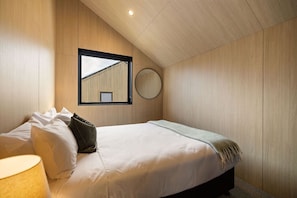 Ein Schlafzimmer mit Bett, Kissen, einer Nachttischlampe, einem runden Spiegel und einem Fenster mit Blick auf ein Gebäude.