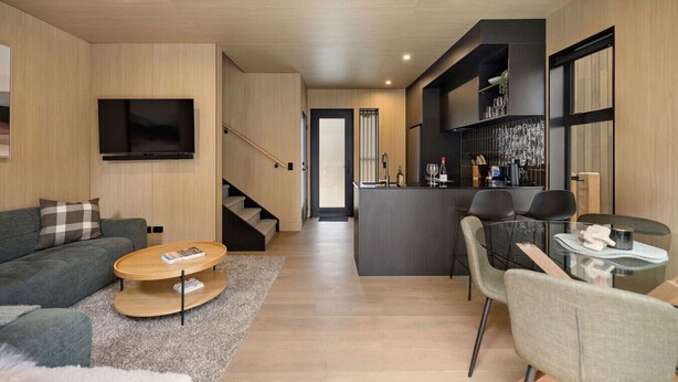 A modern living room with a sofa, coffee table, and TV. A kitchen area with bar stools and a dining table is visible. - Luxury Queenstown 2Bed Villa (Queenstown)