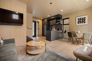 A modern living space with a sofa, a coffee table, a dining area with chairs, a kitchen with a bar, and a flat-screen TV mounted on the wall.