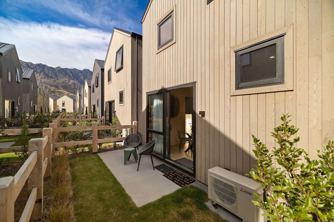 A row of modern wooden buildings with a patio area, outdoor seating, and a view of mountains in the background.