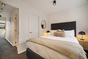 Ein Schlafzimmer mit einem großen Bett, einem dunklen Kopfteil, zwei Nachttischlampen und einer Tür zu einem weiteren Raum.
