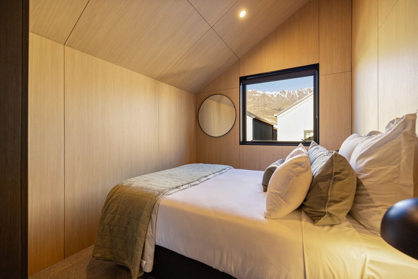 A bedroom with a bed, pillows, a bedside table, a mirror, and a window with a mountain view.