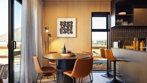 Un espace de salle à manger moderne avec une table ronde, quatre chaises orange, une bouteille de vin et une œuvre d’art abstraite encadrée.