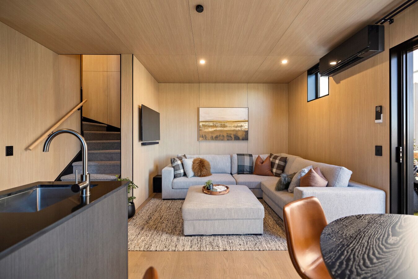 A modern living room with a grey sectional sofa, a coffee table, and a staircase leading to a kitchen area.