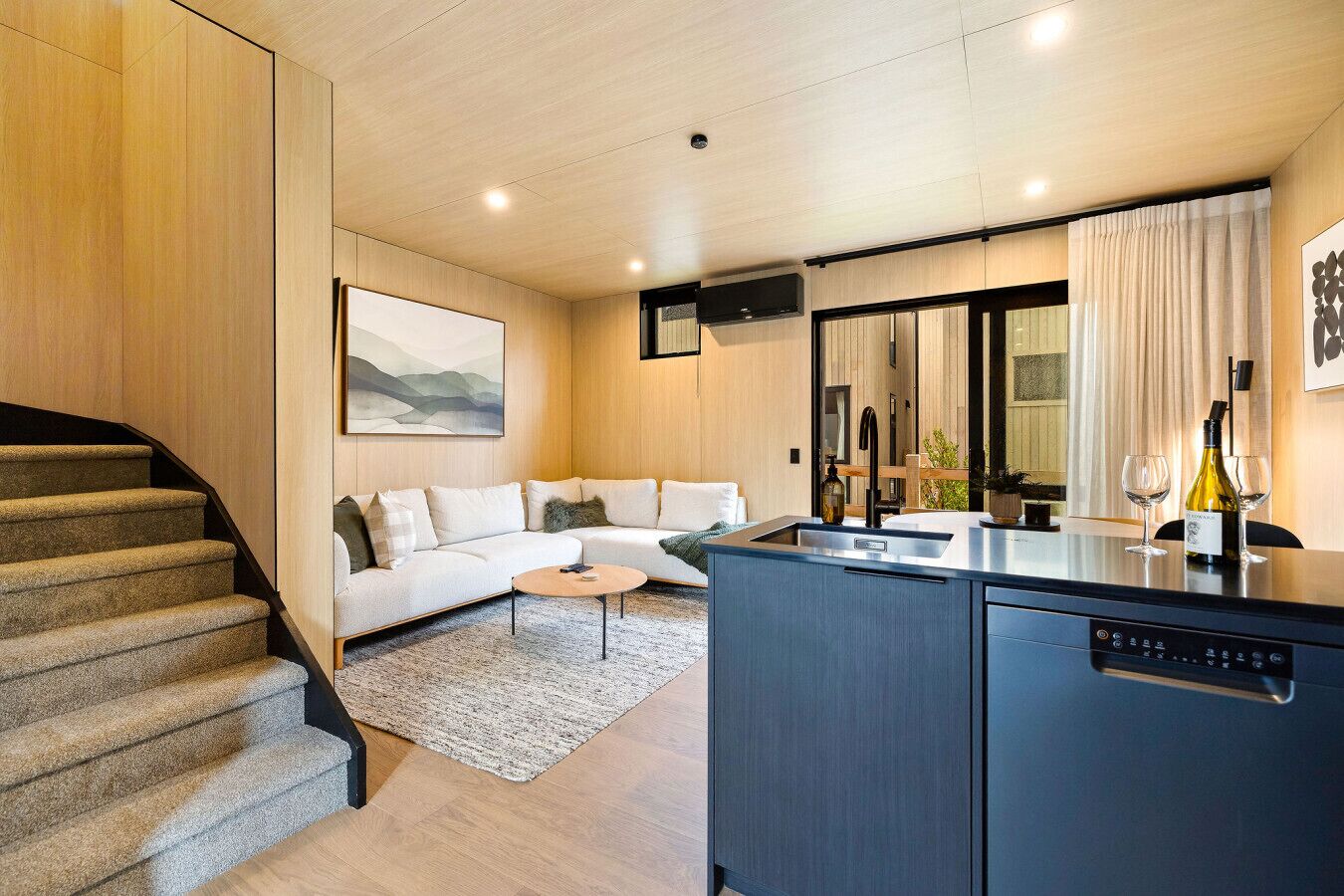 A modern living space with a staircase, a sofa, a coffee table, a kitchen area with a dishwasher, and a dining table with wine glasses.
