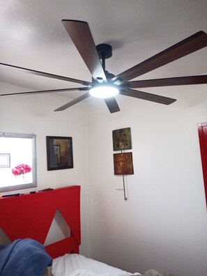 Une pièce dotée d’un ventilateur de plafond, d’un lit, d’une fenêtre donnant sur un jardin fleuri et de deux œuvres d’art accrochées au mur.