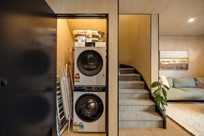 A laundry room with two stacked front-loading washers and dryers, a staircase leading to a living area with a couch and a plant. - Rāua Luxury Villa, Queenstown (Queenstown)