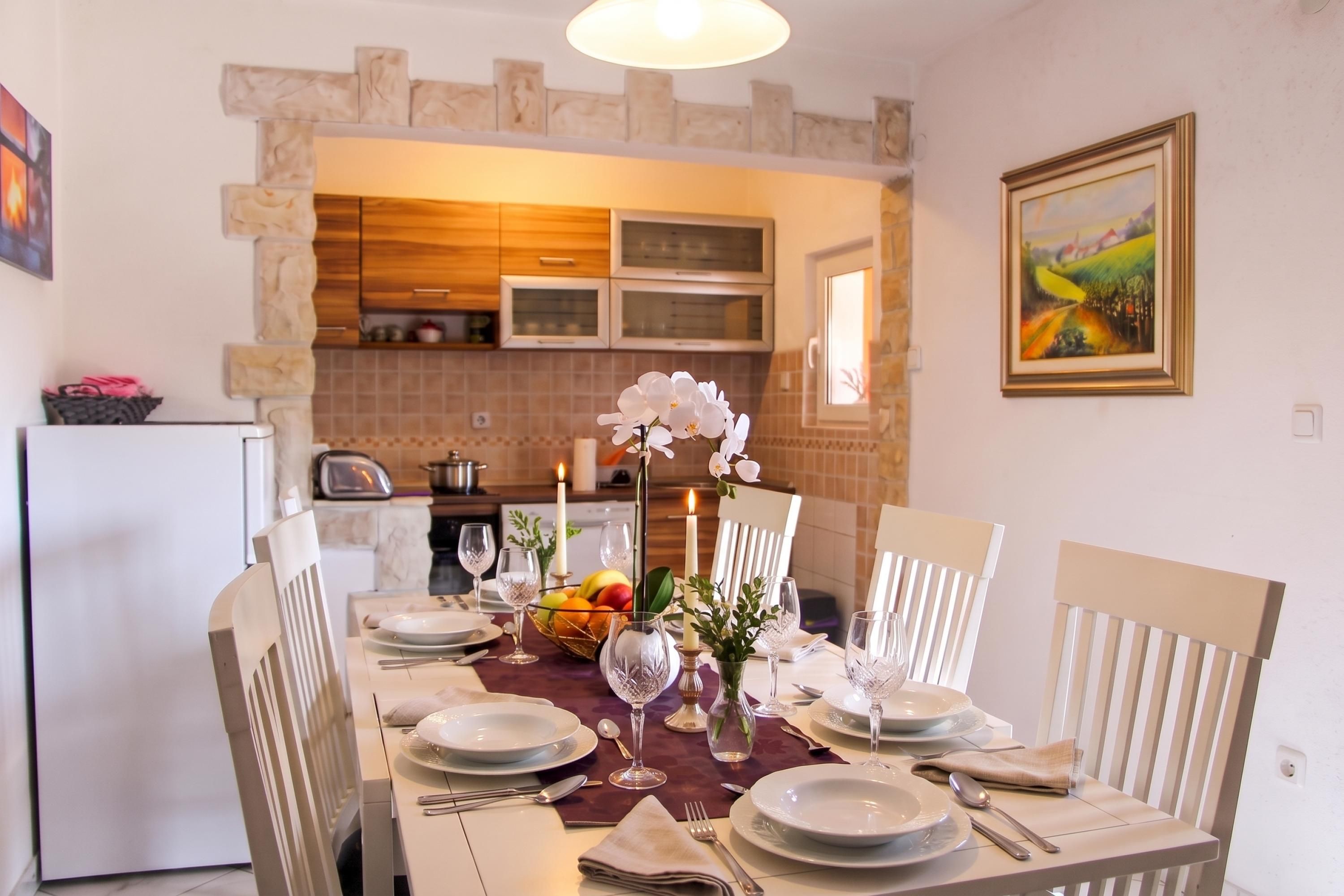 Un coin repas avec une table dressée pour six personnes, des chaises blanches et une cuisine en arrière-plan.