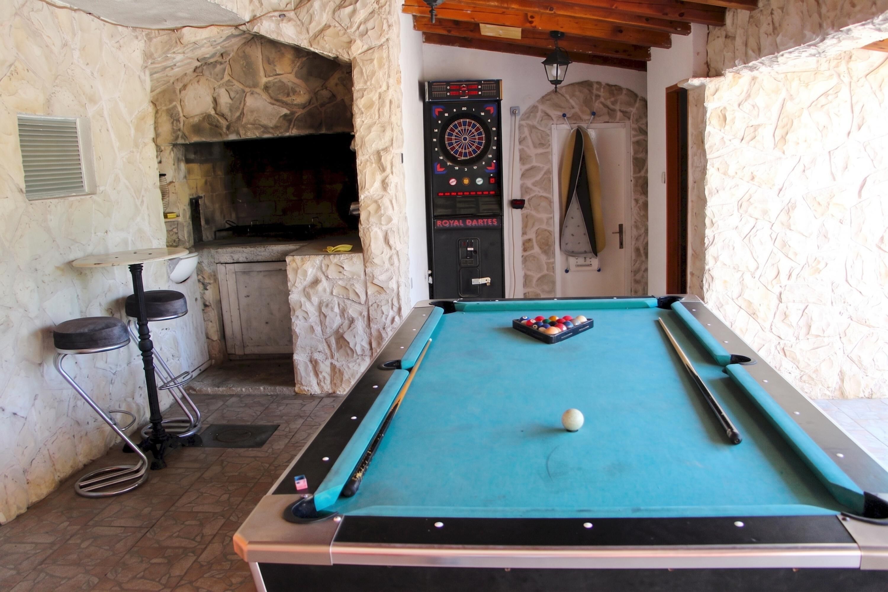 A pool table with a single white cue ball, surrounded by a stone wall and a door leading to a room with a dartboard.