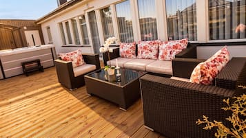 Un espace extérieur aménagé avec des meubles en osier et des coussins à motifs floraux, une table basse en verre et une vue sur un bâtiment doté de grandes fenêtres.
