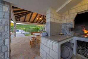 A stone fireplace with a fire, a stone bench, and a wooden table in a covered outdoor area. - Pool Villa Marija, Šibenik, Croatia (Šibenik)