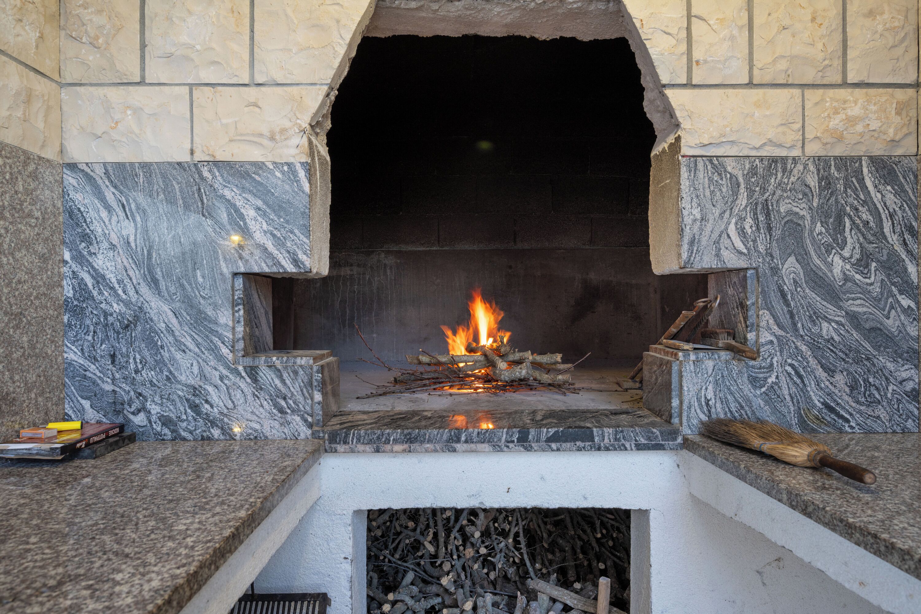 Une cheminée en pierre avec un feu qui crépite, un balai et une pile de bois de chauffage.