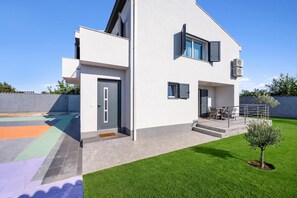Ein modernes, zweistöckiges Haus mit Flachdach, einer Haustür und einer kleinen Veranda mit Sitzgelegenheiten.