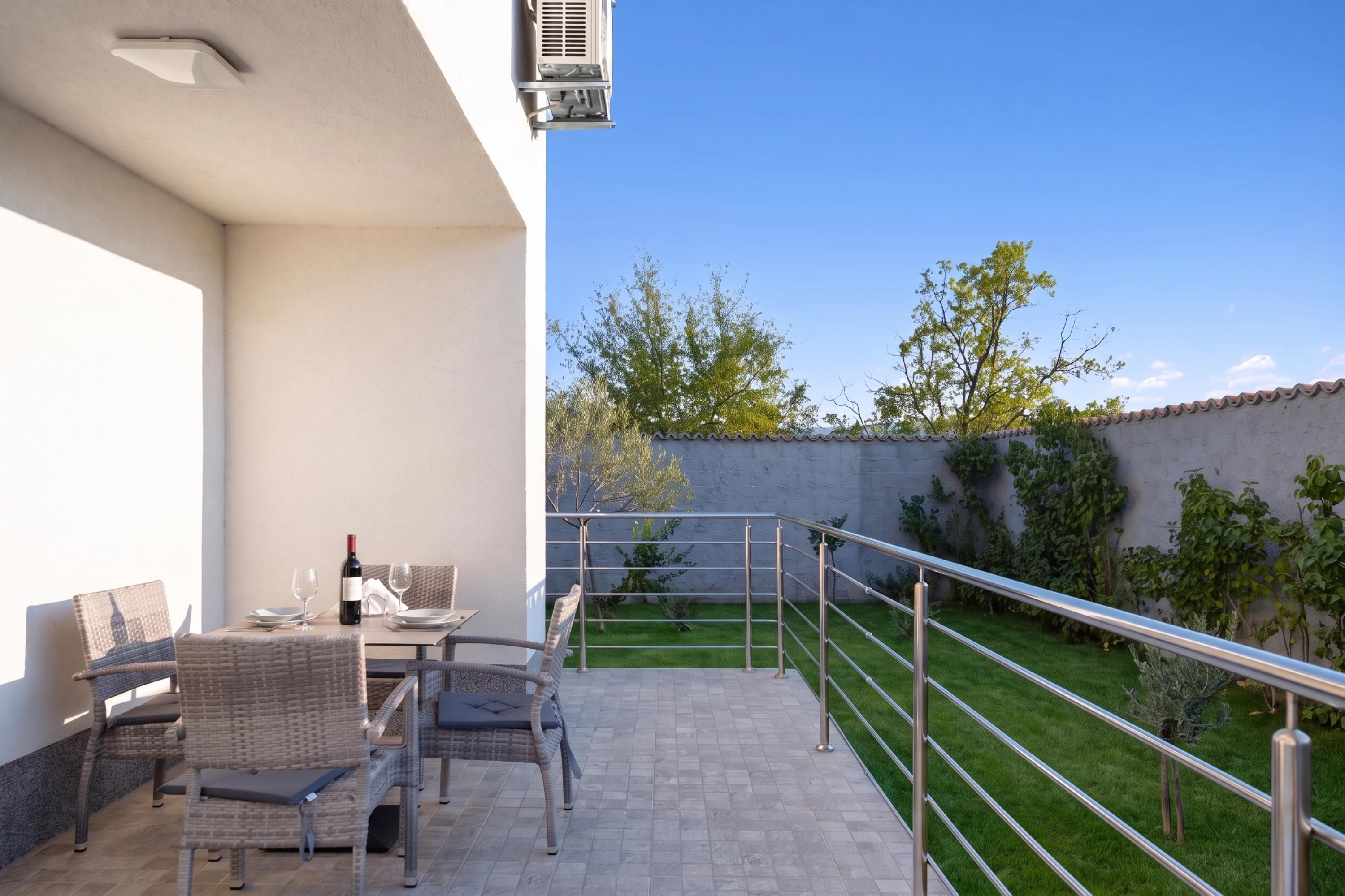 An outdoor patio with wicker furniture, a table with a bottle of wine, and a clear view of a green lawn and trees.