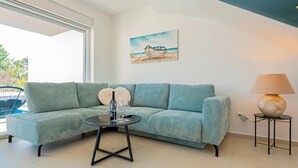 Una sala de estar moderna con un sofá verde, una mesita redonda con una botella de vino y copas, una lámpara y un cuadro de un barco en la pared.