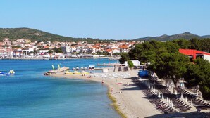 Un pueblo costero con una playa, hileras de sombrillas y una vista despejada del mar y colinas distantes.