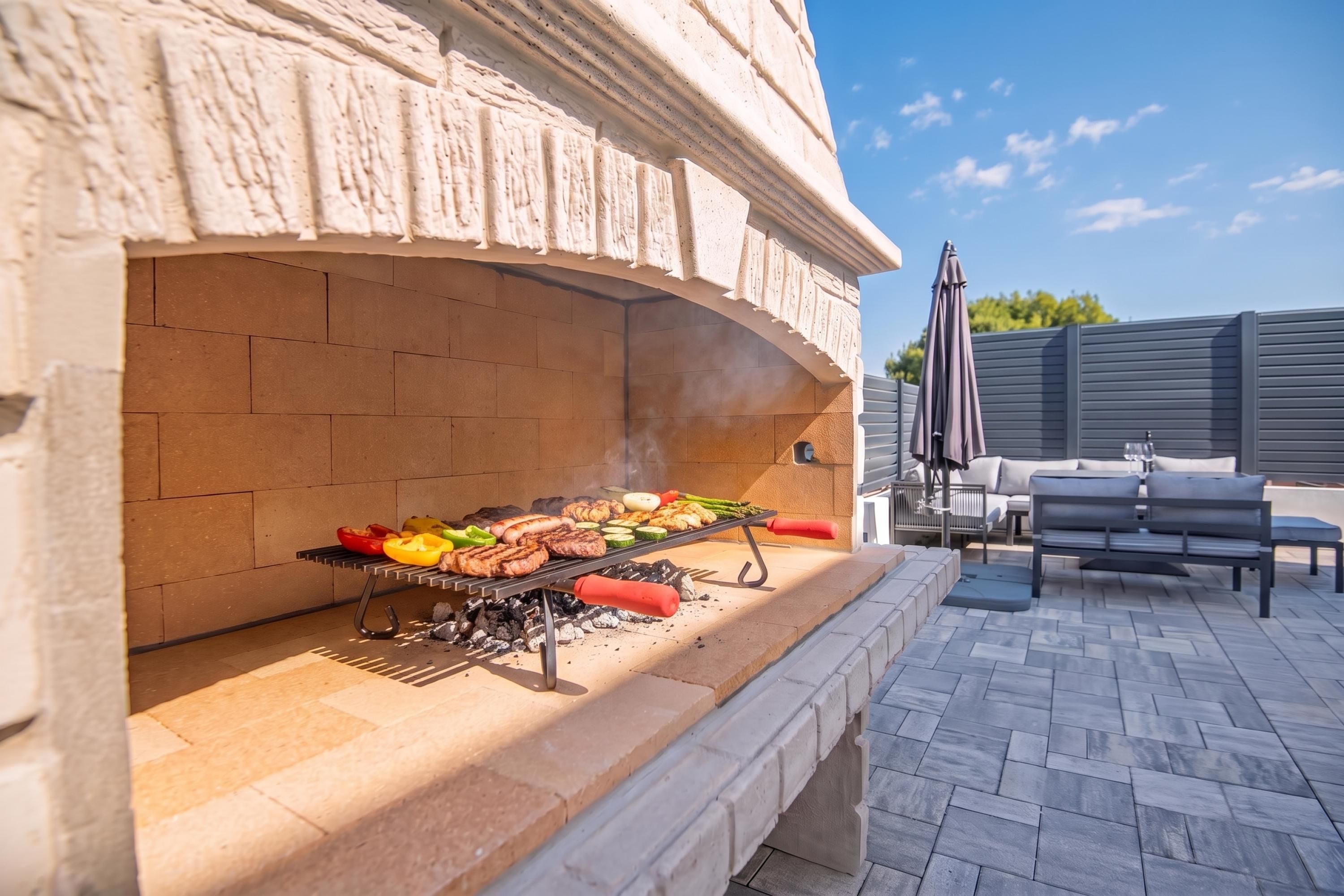 An outdoor grill area with a brick oven, a barbecue grill with various meats and vegetables, and a patio with outdoor furniture.